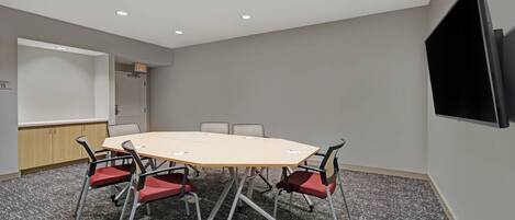 Ein Konferenzraum mit einem großen Tisch, sechs Stühlen, einer Whiteboard-Tafel und einem Fernseher an der Wand.