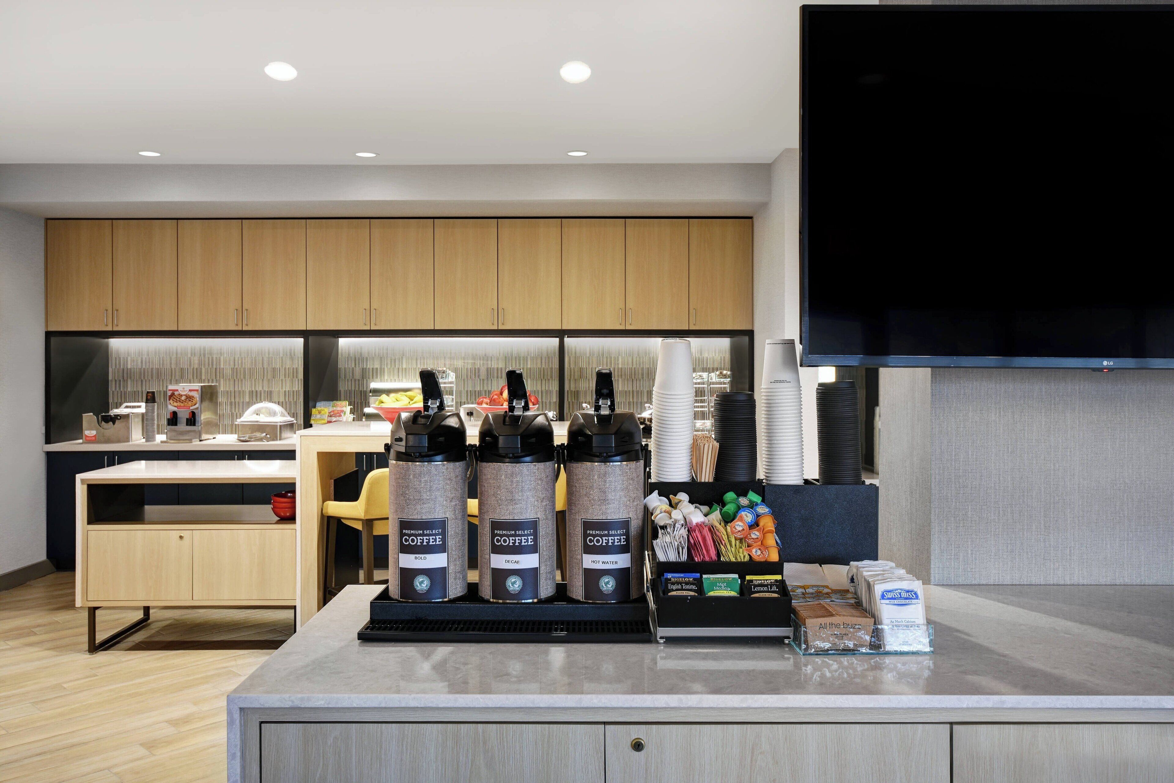 A modern coffee station with three coffee dispensers, a counter with various coffee products, and a television mounted on the wall.
