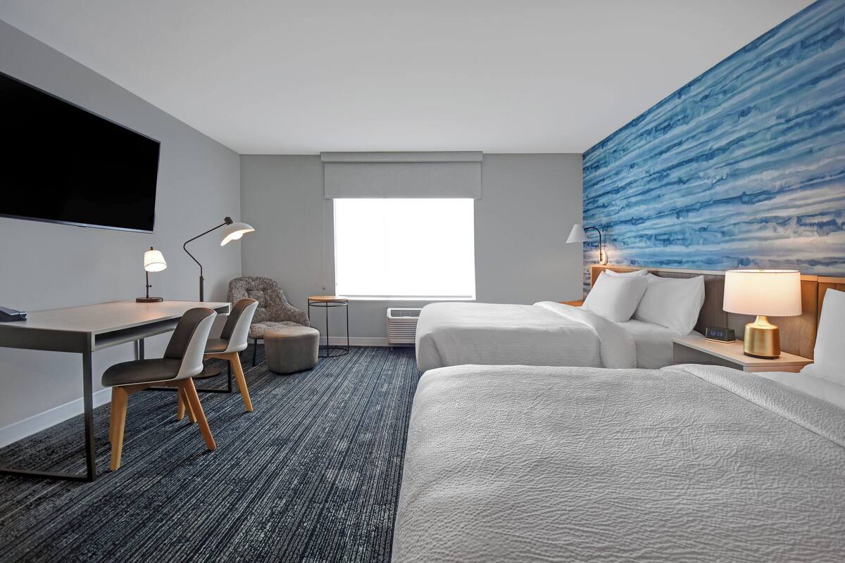 A hotel room with a desk, two beds, a large window, a flat-screen TV, and a blue textured wall.