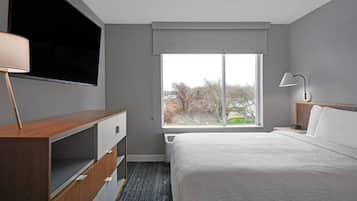 Een hotelkamer met een bed, een aan de muur bevestigde televisie, een nachtkastje met een lamp en een raam met uitzicht op bomen.