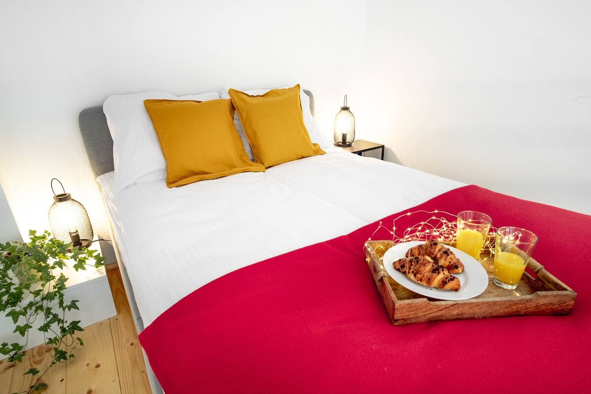 Un lit bien fait, avec des oreillers blancs et moutarde, une couverture rouge et un plateau de petit-déjeuner contenant des croissants et du jus d’orange.
