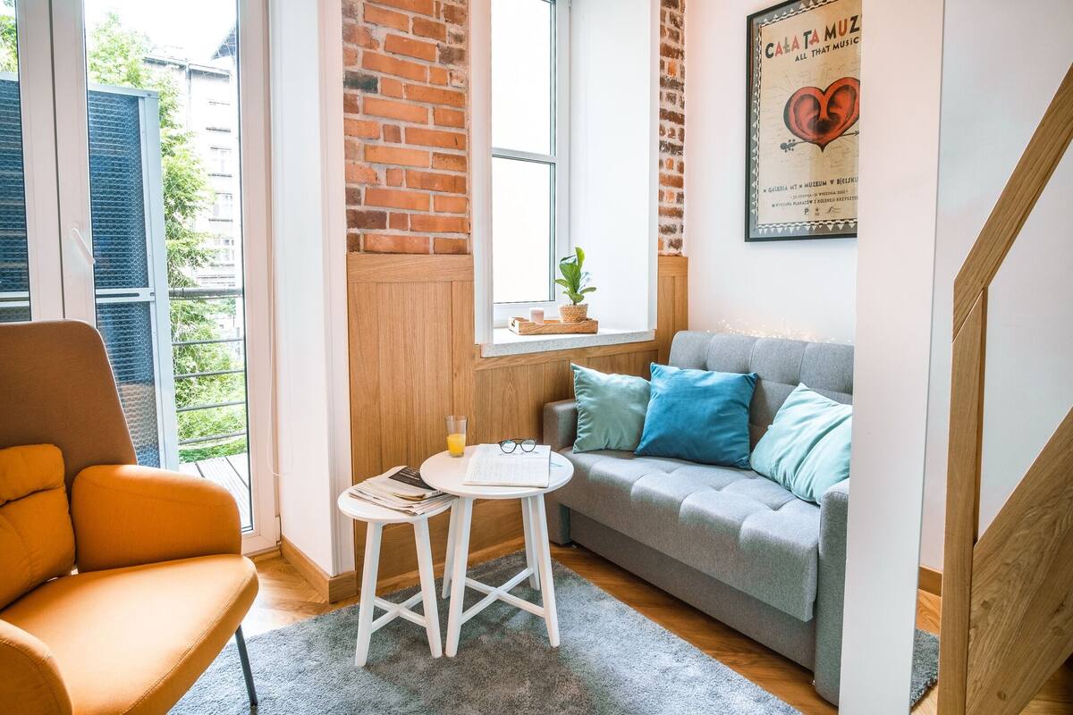Un salon chaleureux avec un canapé gris, un fauteuil orange et une petite table blanche sur laquelle se trouvent un livre et des lunettes.