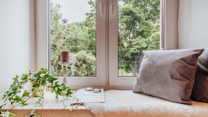 Un coin lecture confortable avec un banc rembourré, une petite table sur laquelle est posée une bougie, et une fenêtre donnant sur un paysage verdoyant.