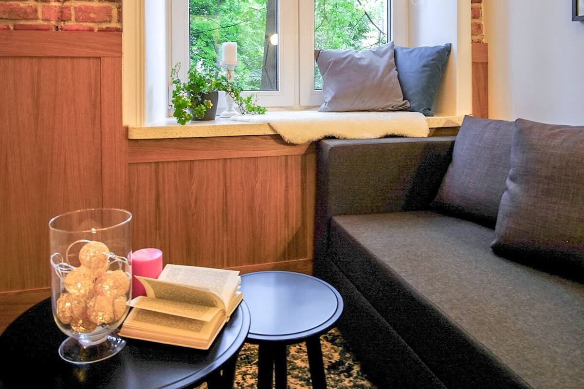 Une pièce chaleureuse avec un mur en briques, une fenêtre donnant sur les arbres, un canapé, une petite table avec un livre et une bougie, et un saladier en verre rempli de biscuits.