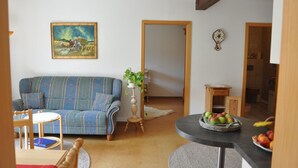 A living room with a blue sofa, a small round table, a wooden chair, a painting on the wall, and a basket of fruit on a counter. - Ferienwohnung Gäste Oase (Bad Windsheim)
