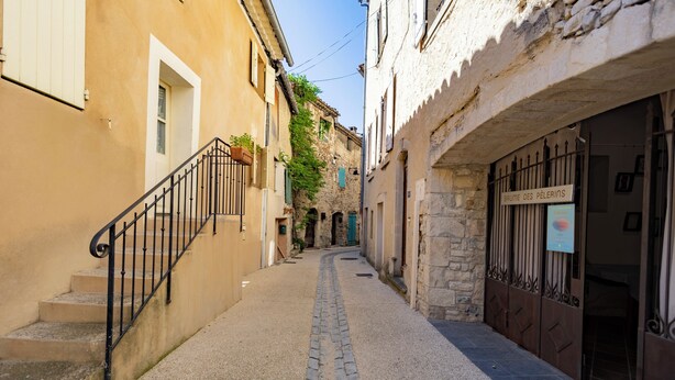 A narrow cobblestone street flanked by buildings with stone facades and metal gates. - La Baume des Pèlerins (Sablet)