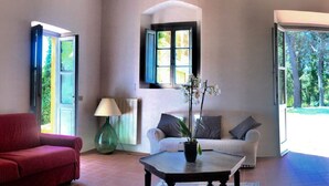 Ein Wohnzimmer mit einem roten Sofa, einem weißen Sofa, einem Couchtisch und einer Lampe. Es gibt zwei Fenster und eine Tür.
