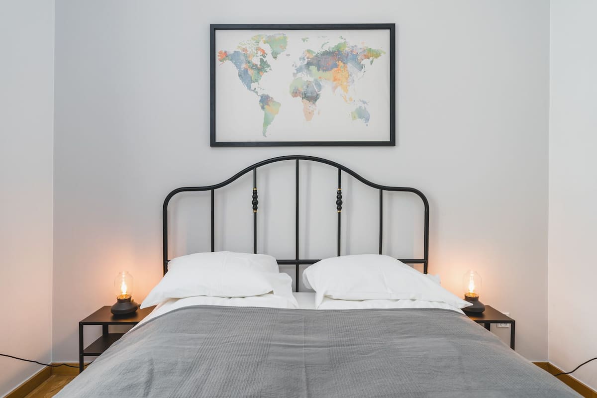 Uma cama bem arrumada com um cobertor cinza, duas almofadas brancas e uma cabeceira de metal preta. Há duas mesas de cabeceira com abajures, e um mapa-múndi emoldurado está pendurado na parede acima da cama.