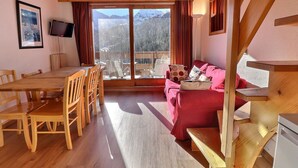 A room with a wooden dining table, chairs, a red sofa, a TV on the wall, and a large window with a mountain view. - Lovely Quiet Duplex, South Facing, On the Slopes, 2* for 5 People (Méribel)