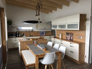 A modern kitchen with wooden cabinets, a central island with seating, and a hanging light fixture. - 06. floating houses (105 m²) Nanna - Floating houses no. 9 with fireplace, sauna and 2 (Kröslin)
