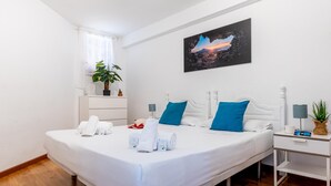 Une chambre d’hôtel avec un grand lit, du linge de lit blanc, un tableau représentant un coucher de soleil, une table de chevet avec une lampe et une plante en pot.