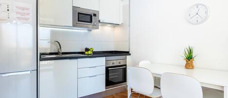 Een moderne keuken met witte kasten, een roestvrijstalen koelkast, een magnetron, een oven en een spoelbak. Er staan twee witte stoelen en een kleine plant op het aanrecht.