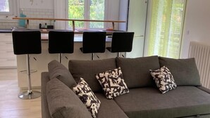 Ein modernes Wohnzimmer mit einem grauen Ecksofa, einem grünen Hocker und einem Esstisch. Im Hintergrund sind drei schwarze Stühle und eine Küchenzeile zu sehen.
