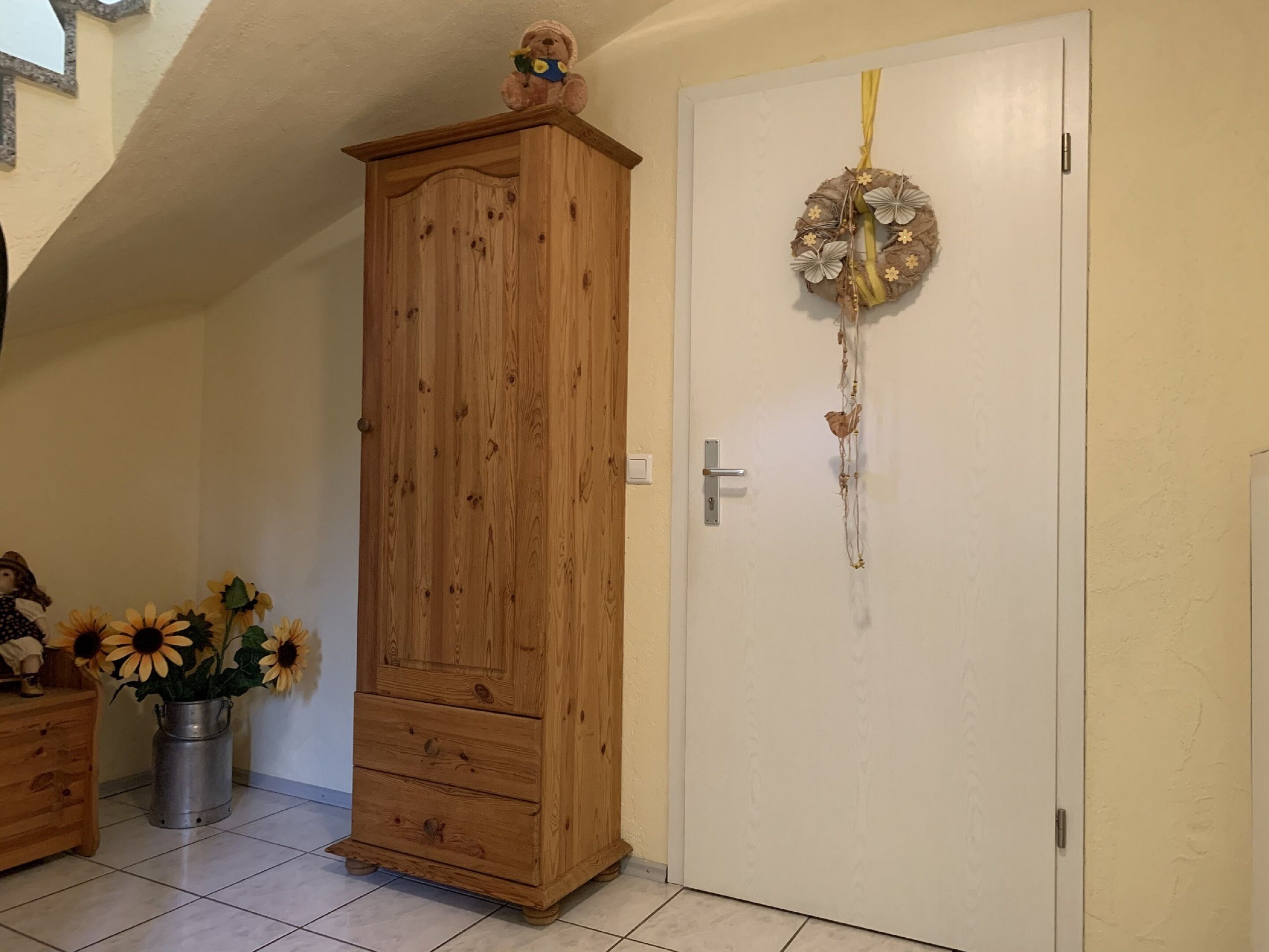 Une armoire en bois surmontée d’un ours en peluche, une porte blanche décorée d’une guirlande et un vase rempli de tournesols posé sur le sol.