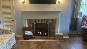 Un salon doté d’une cheminée, d’un téléviseur à écran plat fixé au-dessus et d’une grande fenêtre ornée de rideaux.