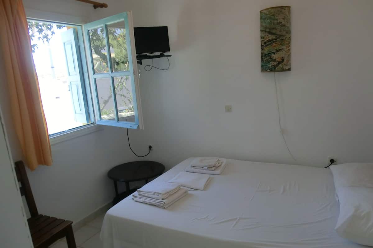 A bedroom with a bed, a small table, a chair, a window, and a wall-mounted TV.