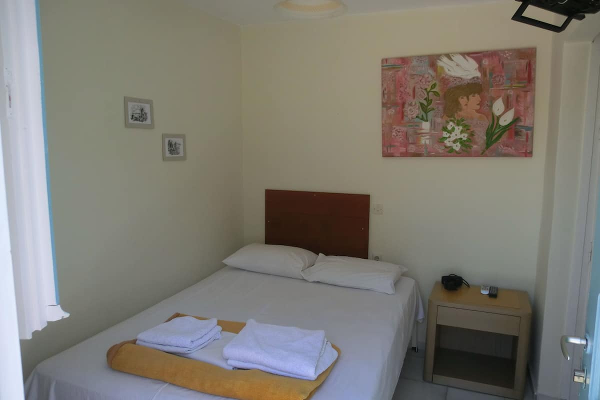 A neatly made bed with white linens and a folded towel, a small bedside table with a remote control, and a painting on the wall.