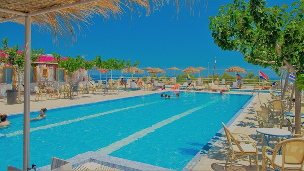A swimming pool with lounge chairs and umbrellas, surrounded by trees and buildings with blue and white facades. - Santorini Camping & Hostel (Santorini)