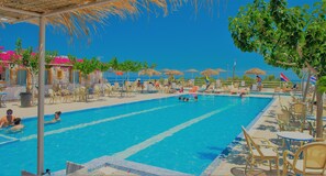 A swimming pool with lounge chairs and umbrellas, surrounded by trees and buildings with blue and white facades. - Santorini Camping & Hostel (Santorini)