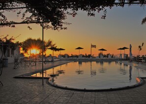 A poolside area with lounge chairs and umbrellas, a flag, and a sunset in the background. - Santorini Camping & Hostel (Santorini)