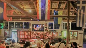 A bar area with multiple TVs, a bar counter, and a variety of bottles. - Santorini Camping & Hostel (Santorini)