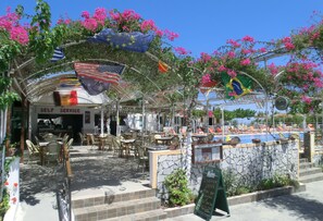An outdoor cafe with tables and chairs, a pool, and flags. - Santorini Camping & Hostel (Santorini)