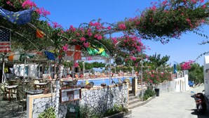 An outdoor café with tables and chairs, a pool, and a stone wall with a signboard. - Santorini Camping & Hostel (Santorini)