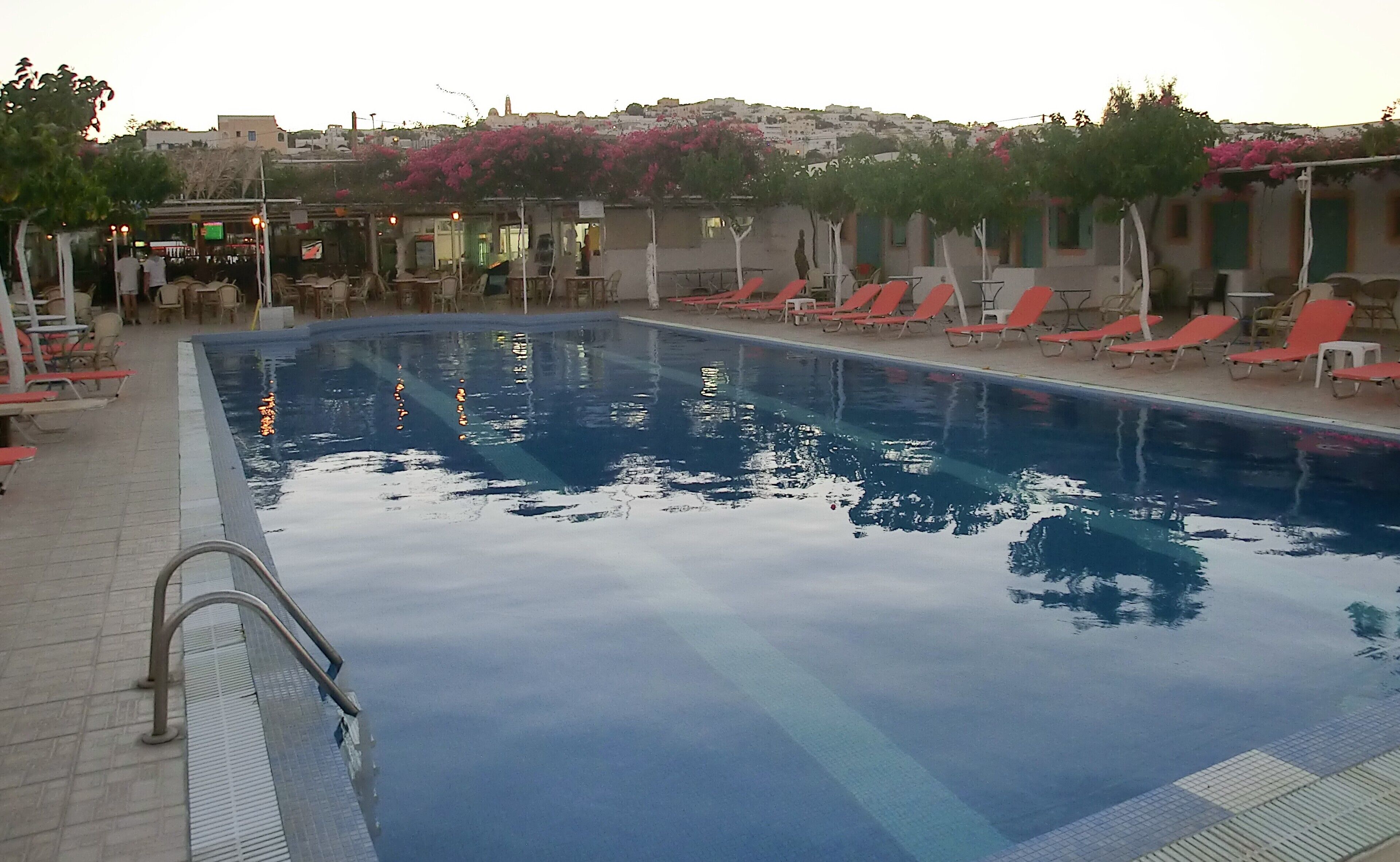 A swimming pool with lounge chairs and tables, surrounded by buildings and pink flowering trees.