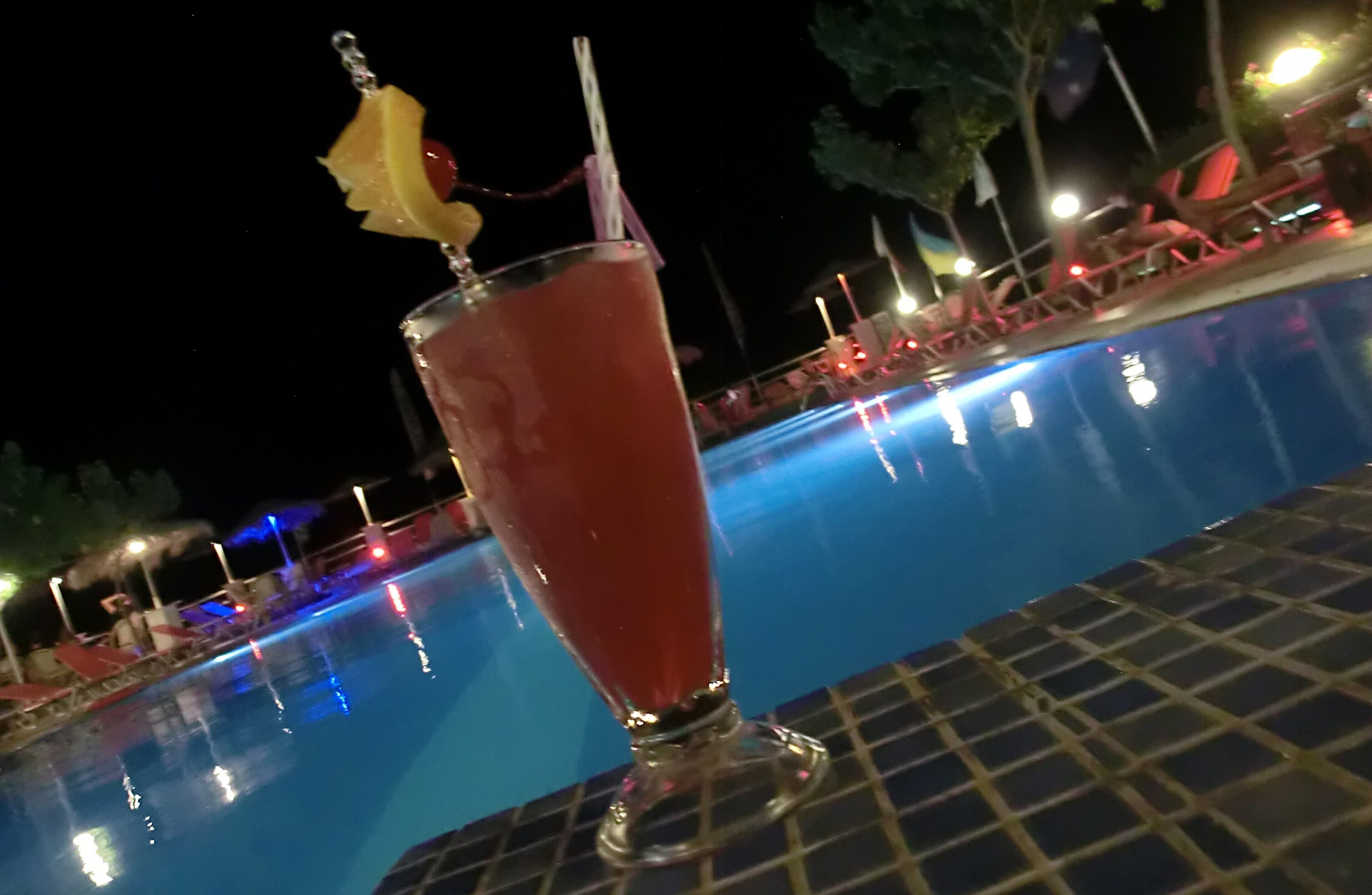 A glass of red beverage with a straw and a cherry on a stick, placed on a tiled surface near a pool.