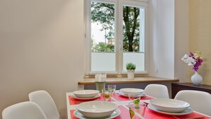 Een eetruimte met een tafel voor vier personen, een raam met uitzicht op bomen en gebouwen, en een bloemenvaas op de vensterbank.