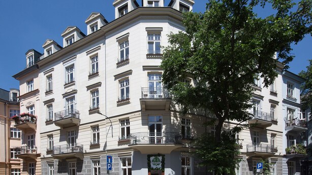 Um edifício com vários andares, varandas, uma entrada visível e uma árvore em frente.
