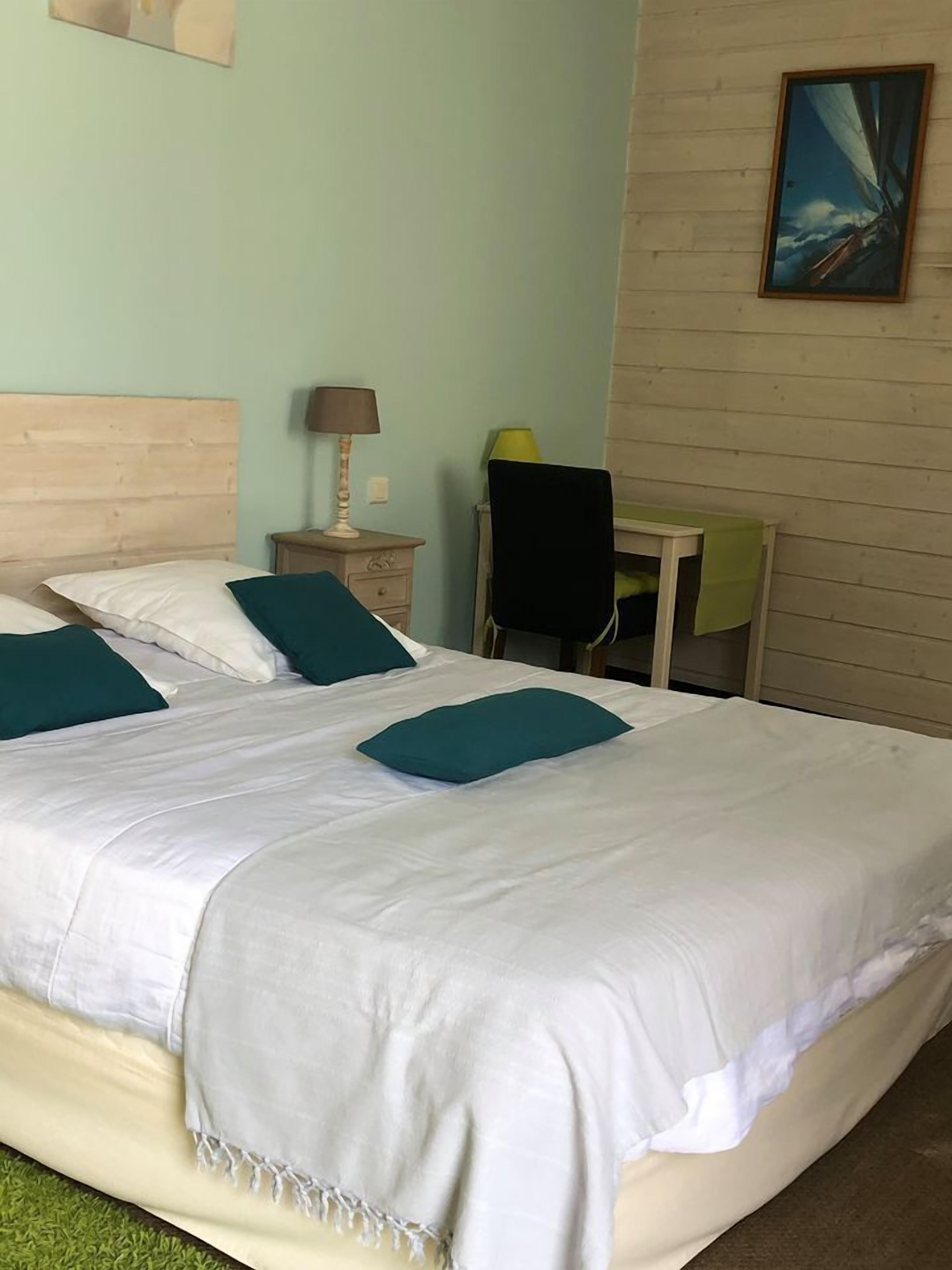 A neatly made bed with white linens and teal pillows, a wooden headboard, a bedside table with a lamp, a chair, and a painting on the wall.