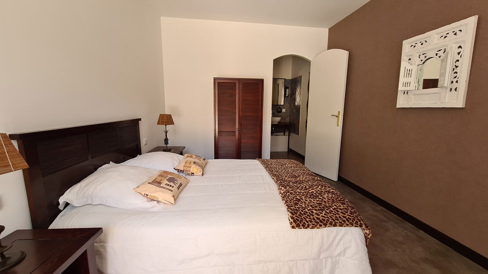 A hotel room with a large bed, a nightstand, a lamp, a wardrobe, and a mirror on the wall.