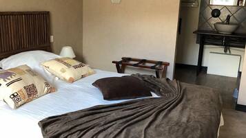Una habitación de hotel con cama, almohadas, una mesita de noche, lámpara, espejo y un pequeño escritorio con lavamanos.