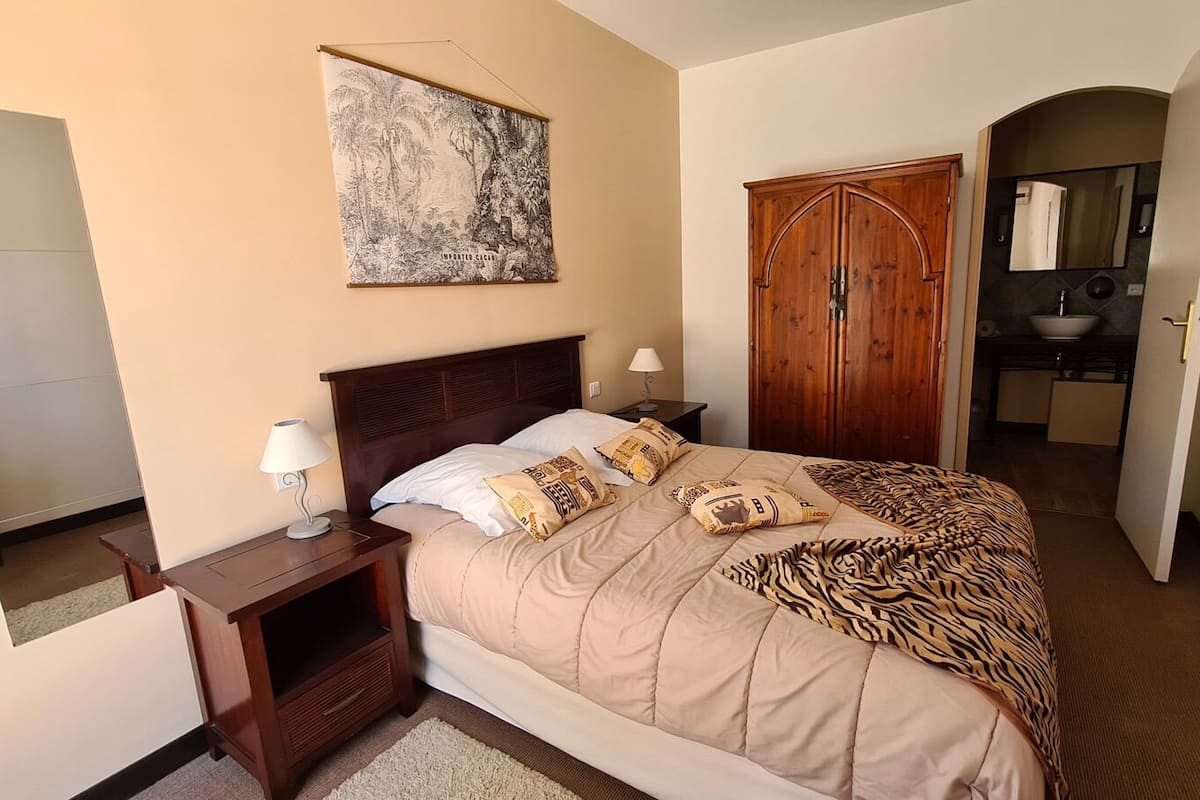 A bedroom with a bed, nightstands, a wooden wardrobe, a bathroom with a sink and mirror, and a wall-mounted artwork.