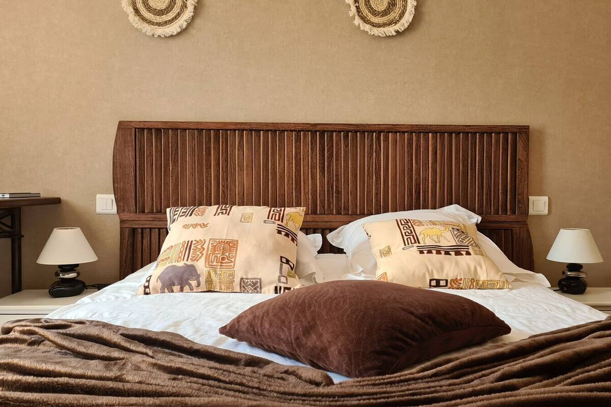 A hotel room with a wooden headboard, a bed with patterned pillows, a brown throw blanket, and bedside lamps.