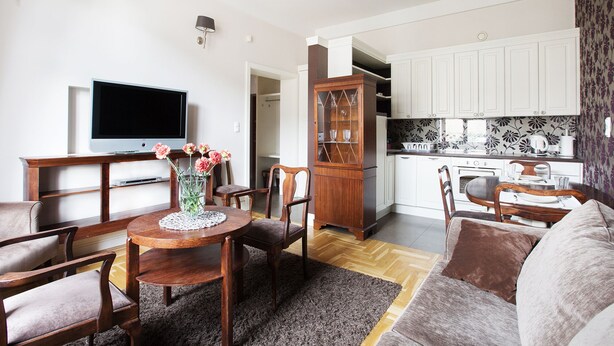 A living room with a TV, dining table, chairs, a vase with flowers, and a kitchen in the background.