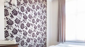 A bedroom with a floral wallpaper, a bed, a nightstand, a lamp, and a window with curtains.