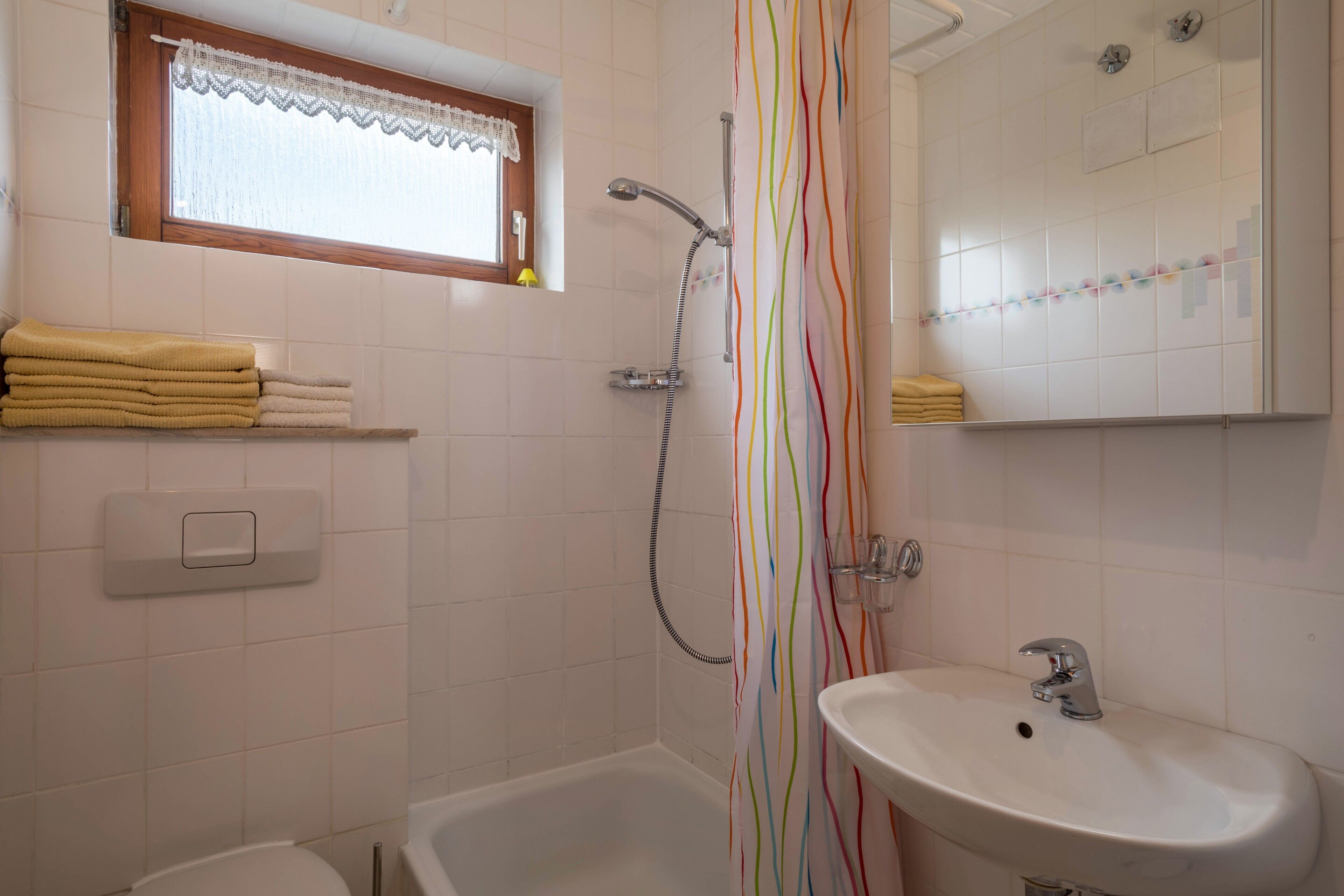Une salle de bain avec une douche, un lavabo, une fenêtre ornée d’un rideau en dentelle et un porte-serviettes.