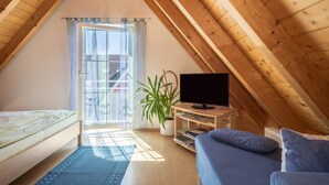 Um quarto com teto inclinado, uma cama, um sofá, uma televisão, uma planta e uma janela com cortinas.