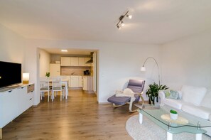 Un salon moderne avec un téléviseur à écran plat, une table basse en verre, un canapé blanc, un coin repas avec des chaises blanches et une cuisine en arrière-plan.