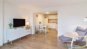 Ein modernes Wohnzimmer mit Flachbildfernseher, Essbereich mit Stühlen, einer Küche im Hintergrund und einem Relaxsessel.