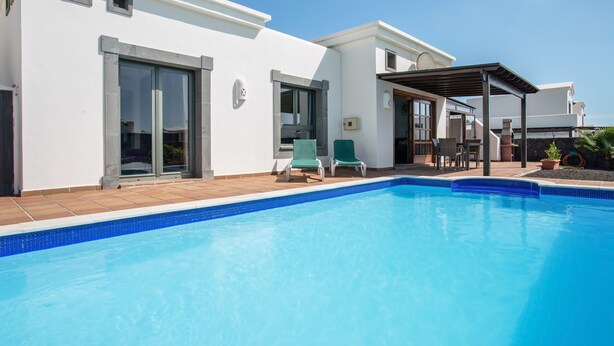 A modern poolside area with a clear blue pool, a covered seating area, and a white building with large windows. - Beautiful Holiday Home “Casa Violeta” with Terraces and Pool (Playa Blanca)