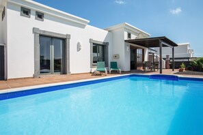 A modern poolside area with a clear blue pool, a covered seating area, and a white building with large windows. - Beautiful Holiday Home “Casa Violeta” with Terraces and Pool (Playa Blanca)