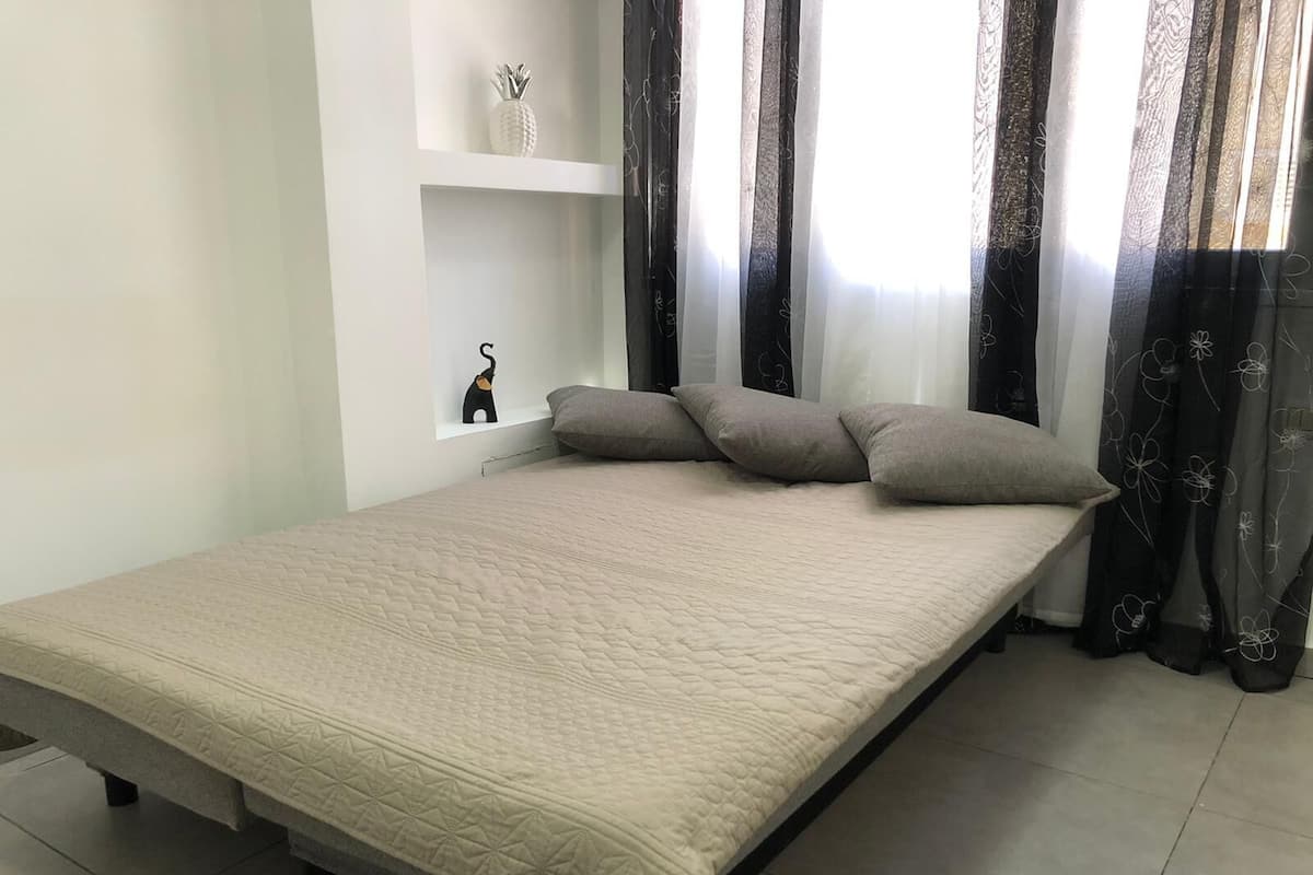 Ein ordentlich bezogenes Bett mit einer gesteppten Decke, eine kleine Ablage mit Dekoration und ein Fenster mit durchsichtigen Vorhängen.