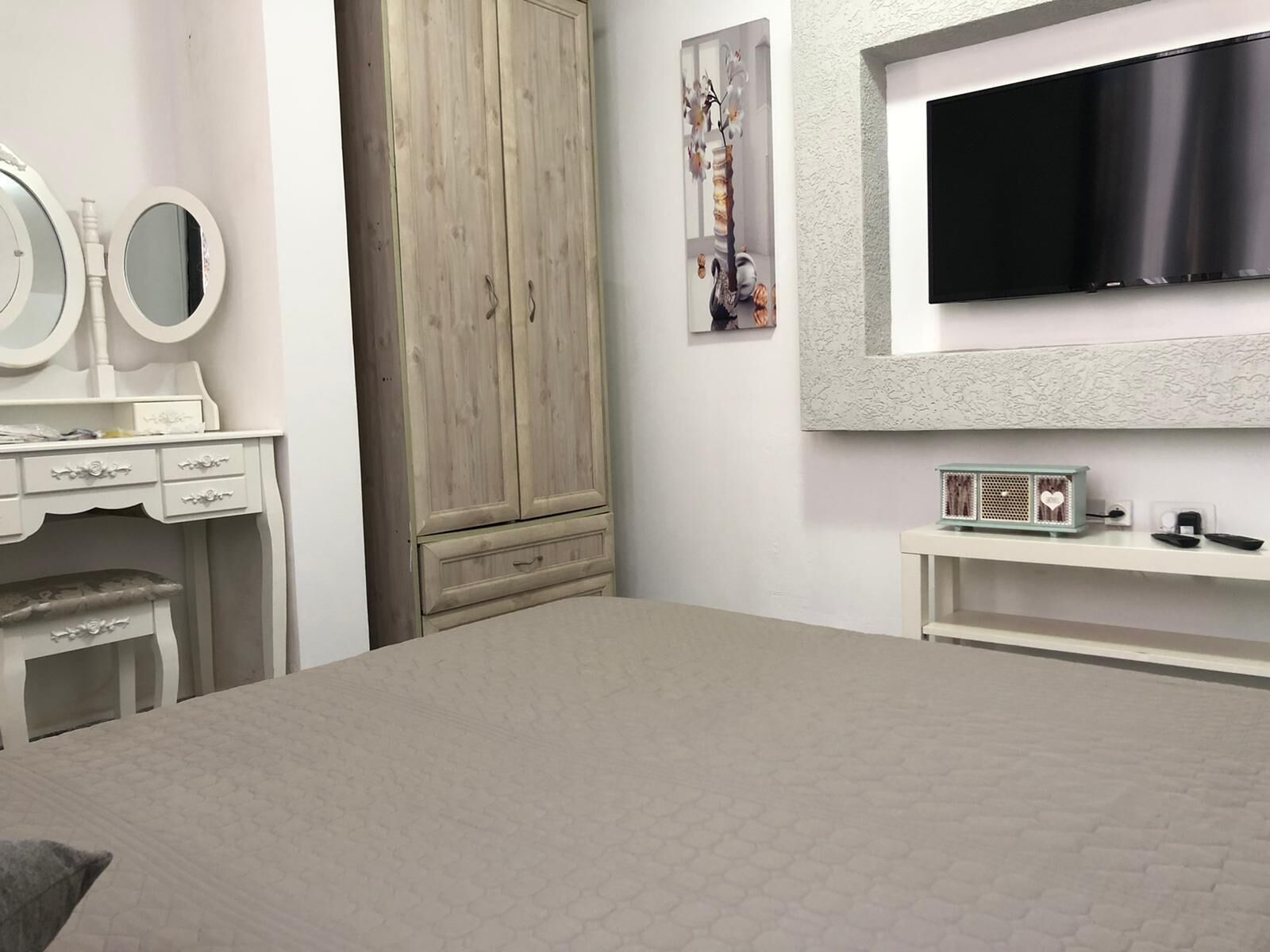 Een slaapkamer met een bed, een dressoir met spiegel, een kledingkast en een aan de muur bevestigde televisie.
