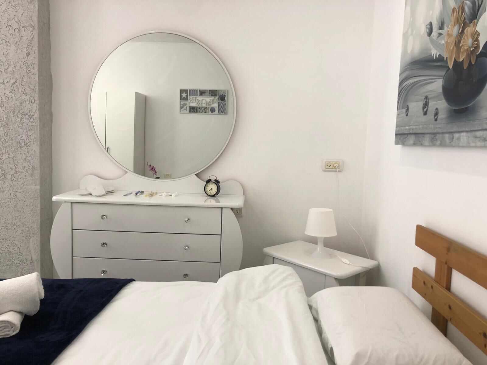 A bedroom with a bed, a dresser with a mirror, a bedside table with a lamp, and a wall-mounted power outlet.