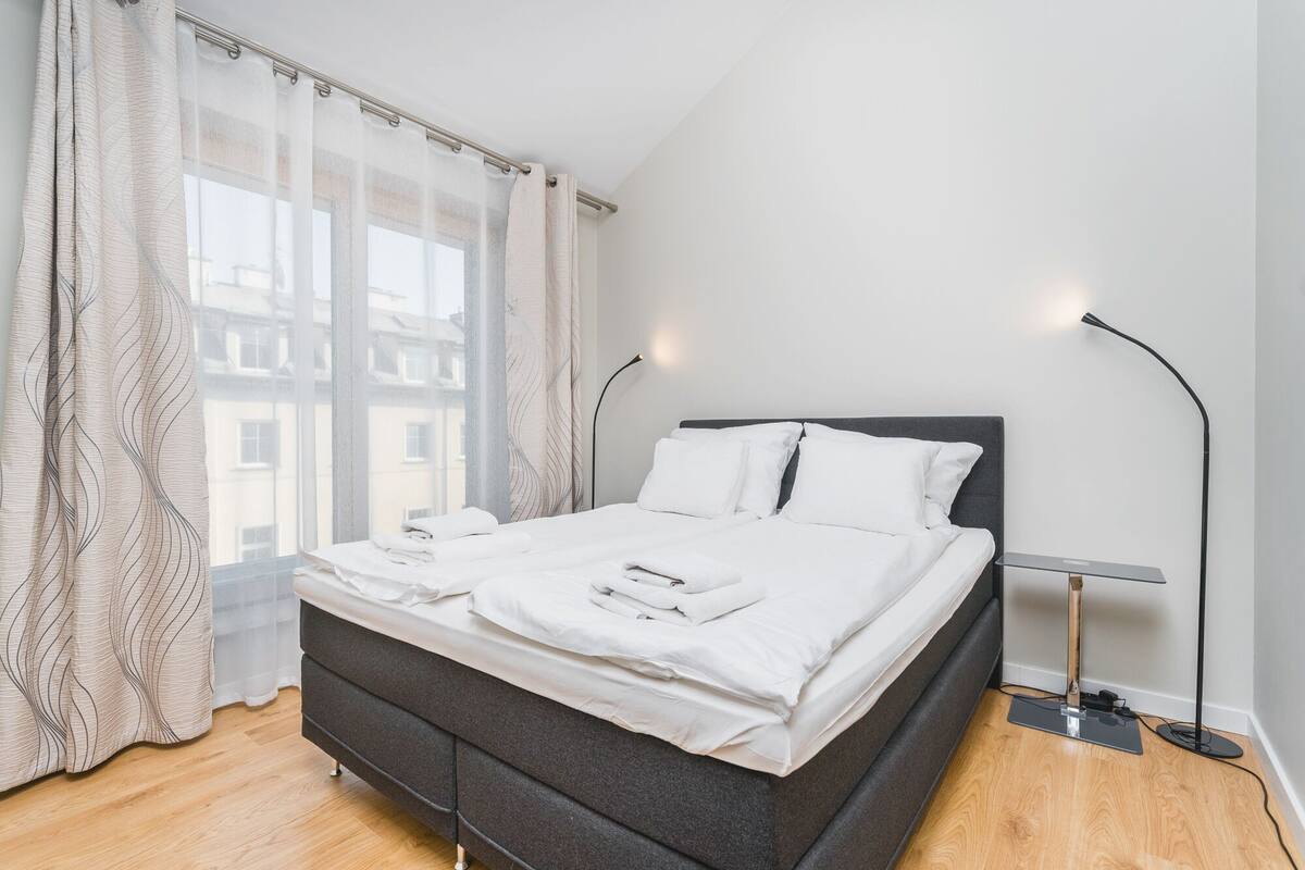 Un lit bien fait, avec du linge de lit et des serviettes blanches, une tête de lit gris foncé et deux lampes de chevet.
