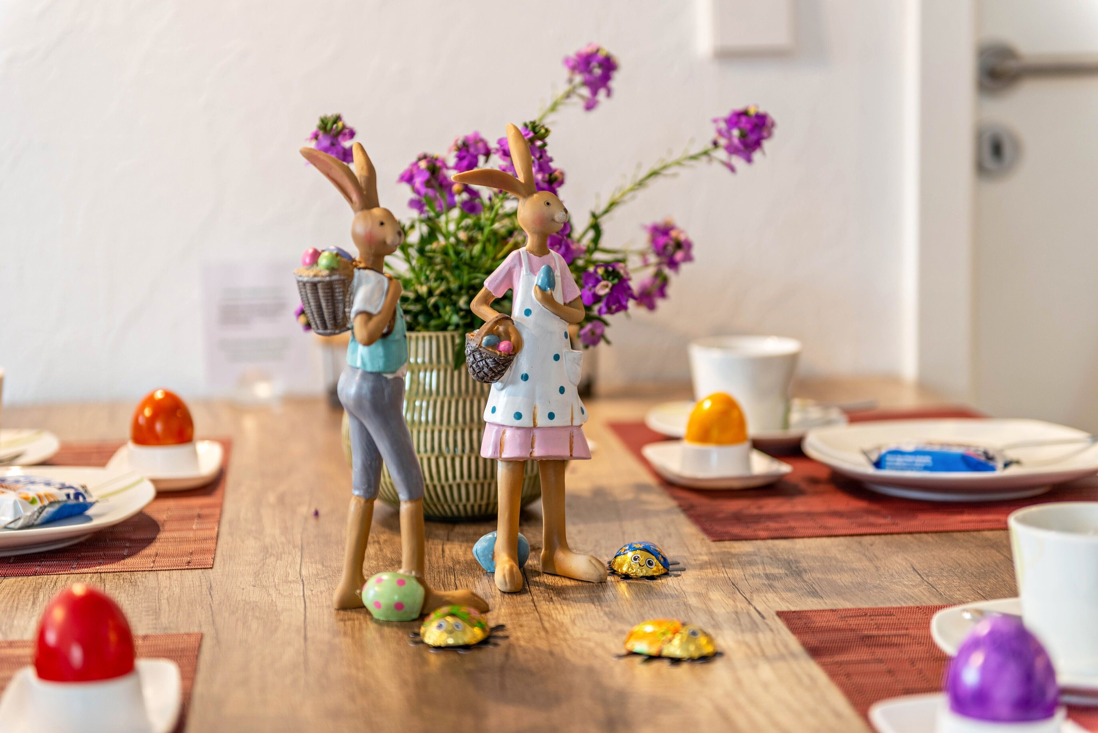 Ein Esstisch mit Keramik-Hasenfiguren, einer Vase mit lila Blumen und verschiedenen Speisen.