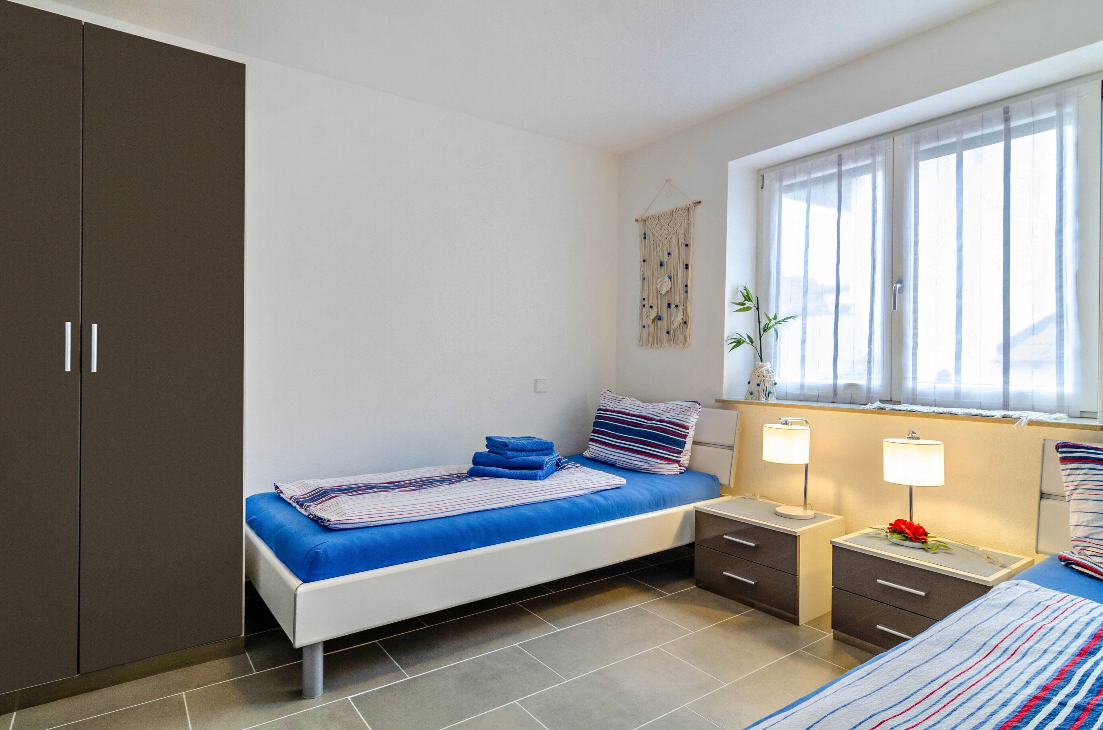 Ein Schlafzimmer mit einem Bett, Nachttischen, einem Kleiderschrank, einem Fenster mit Vorhängen und einem Wandbehang aus Makramee.